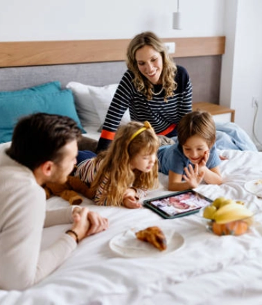Family enjoying breakfast and screen time together on a hotel bed during a staycation.