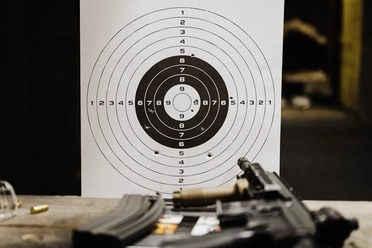 Target board at an indoor shooting range with bullet holes and a firearm in the foreground.