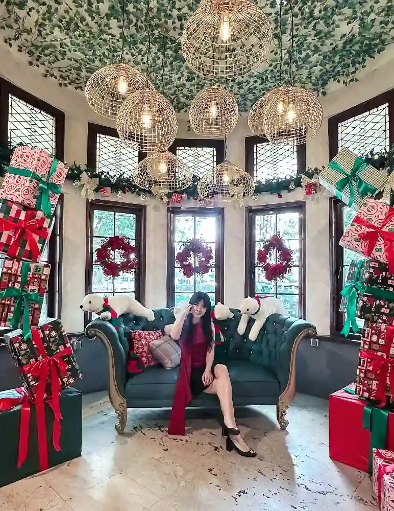 A woman sits on a teal sofa in a Christmas-themed corner decorated with gift boxes, wreaths, and polar bear plushies.