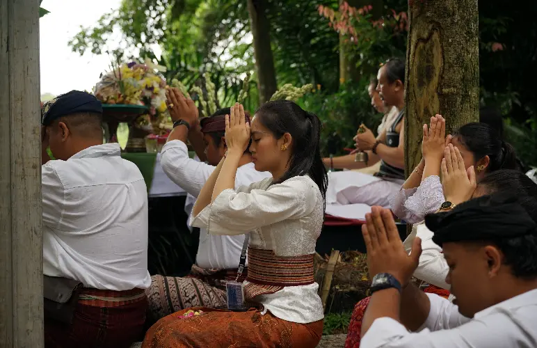 A group of people dressed in traditional Balinese attire sitting outdoors in prayer, hands pressed together, during a ceremonial ritual.